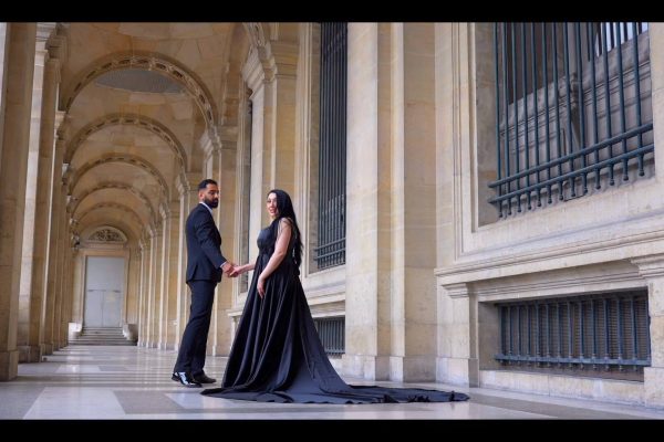 A couple dressed elegantly in black standing in a grand architectural hallway.